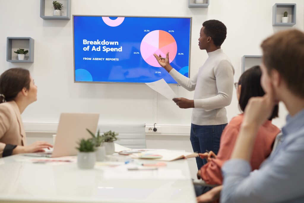 The image shows a professional meeting where a businessman is giving a presentation on ad spend and their PPC Serives. He stands in front of a screen displaying a colorful pie chart labeled "Breakdown of Ad Spend from Agency Reports," explaining the data to colleagues seated around a conference table. The setting appears to be a modern, well-lit office, with the audience paying close attention to the presentation